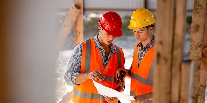 Two men dressed in shirts, orange work vests and helmets explore construction documentation on the building site near the wooden building